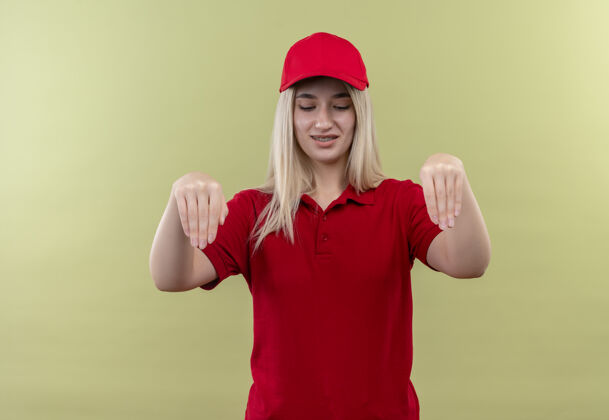 红色看下交付的年轻女孩穿着红色t恤和戴着牙套帽子显示采取在孤立的绿色背景的姿态年轻送货T恤