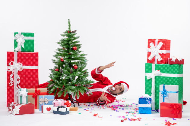 圣诞老人喜庆的节日气氛与年轻的圣诞老人躺在圣诞树后 靠近礼物的白色背景图片圣诞礼物年轻