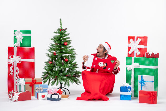 圣诞树新年气氛与有趣的惊喜圣诞老人坐在地上 并显示圣诞袜子附近的礼物和装饰圣诞树上的白色背景装饰圣诞老人坐着