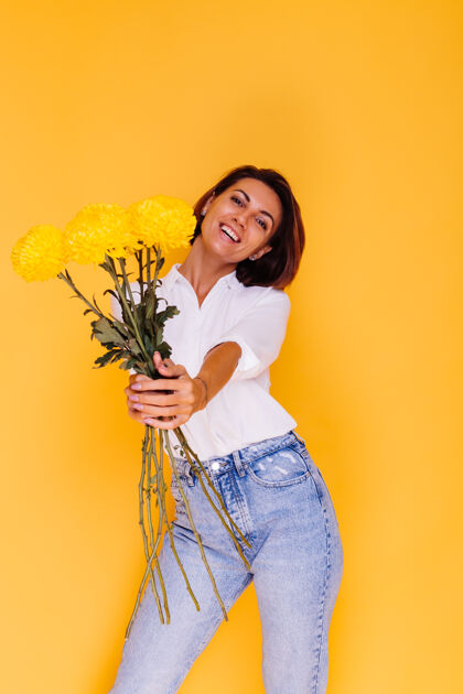 积极摄影棚拍摄的黄色背景快乐的白人妇女短发穿着休闲服白衬衫和牛仔裤手持一束黄色紫苑植物惊喜期待