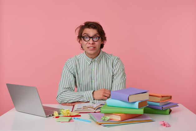 野性一幅年轻人的画像 黑发做鬼脸 拿着书和笔记本电脑坐在工作台上 双手交叉放在台面上表情长相20多岁