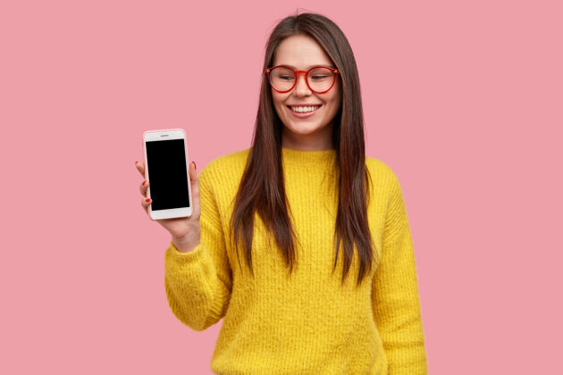 示范乐观的黑发女士展示智能手机屏幕 乐于购买新产品 戴眼镜和黄色毛衣电话展示保持