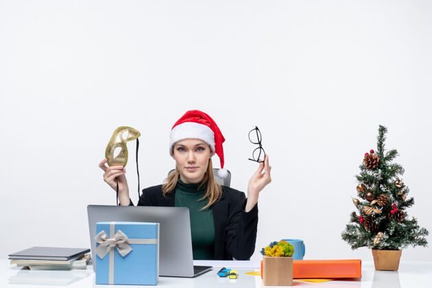 美女在白色背景的办公室里 一位好奇的美女戴着圣诞老人帽 眼镜和面具坐在一张桌子旁 桌子上放着圣诞树和礼物坐着笔记本好奇