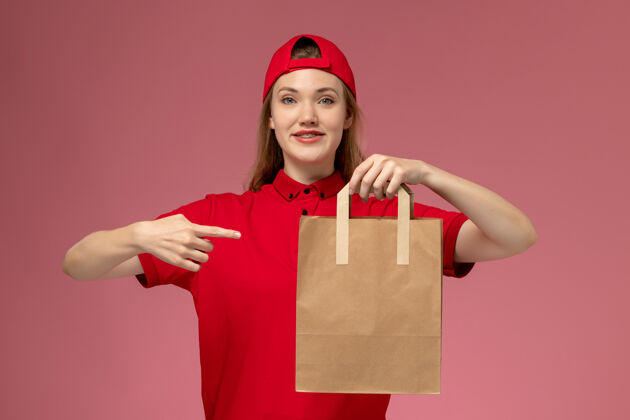 风景正面图：身穿红色制服的年轻女快递员拿着快递纸食品包在浅粉色的墙上食物包裹漂亮