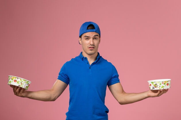 人正面图：身穿蓝色制服披肩的年轻男性信使手持圆形的投递碗站在浅粉色的墙上服务视图成人