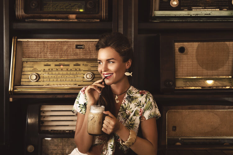 收音机年轻快乐的女人在时尚复古的咖啡馆里喝着冰咖啡 里面有很多老式收音机爱时尚旧的