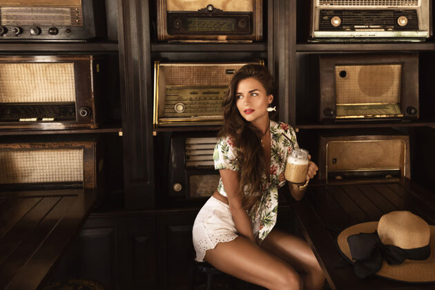 模型年轻快乐的女人在时尚复古的咖啡馆里喝着冰咖啡 里面有很多老式收音机化妆品酒杯时尚