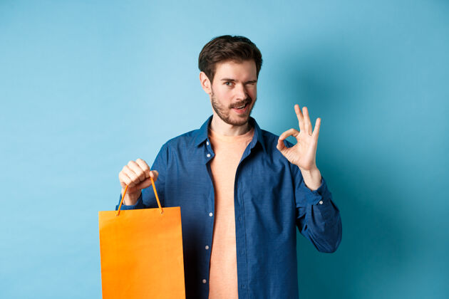 商店穿着休闲装的现代男人展示购物袋和OK标志 对着镜头眨眼 推荐商店 站在蓝色背景上购物者自信人