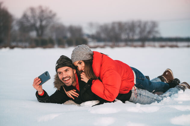快乐一对穿着暖和衣服的快乐的年轻夫妇一起躺在雪地上 一边用智能手机自拍 一边在冬天的乡间玩耍雪喜欢一起