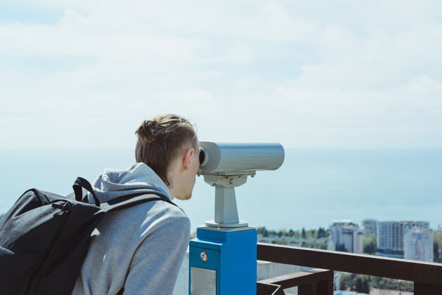 操作年轻的潮人旅游者透过金属硬币操作的望远镜观看海 天和城市 横向生活方式股票图片游客冒险观光