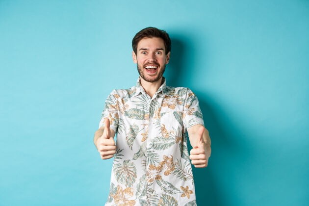 休闲穿着夏威夷衬衫的兴奋的微笑着的家伙竖起大拇指 看起来很惊讶 看看酷的宣传片 站在蓝色的背景上男人酷成人