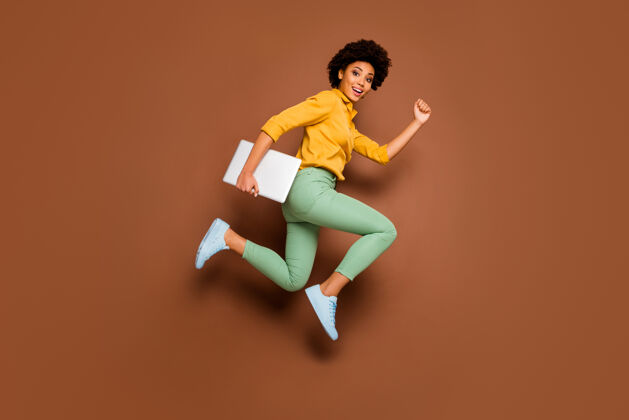 女性时髦深色皮肤女士跳高持笔记本抓紧上课上课女生穿黄色衬衫绿色裤子鞋孤立的棕色微笑跑步飞行