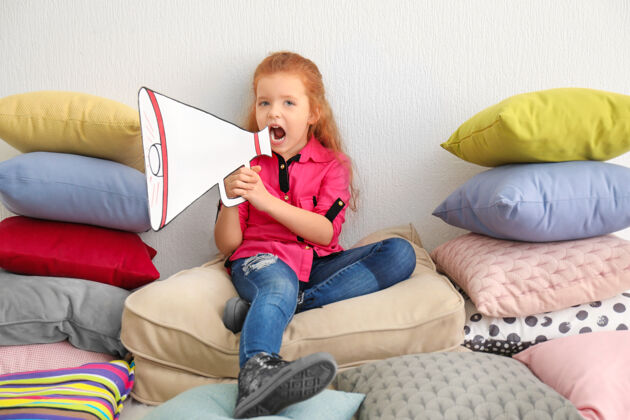 活动可爱的小女孩和纸扩音器坐在一堆枕头在室内讲话可爱扩音器