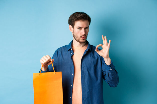 脸帅哥推荐店 展示橙色购物袋和ok标志 站在蓝色背景上肖像年轻购物