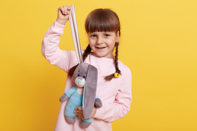 玩耍微笑的小女孩儿拉着毛茸茸的小兔子的耳朵 看着相机和快乐的表情 孩子在淡粉色衬衫摆出孤立的黄色背景孩子兔子天真
