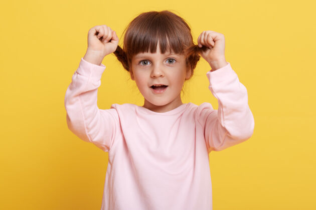 毛衣快乐的小女孩扎着辫子 张着嘴直视镜头 手放在辫子上 穿着淡粉色的休闲装 在黄色背景下摆出孤立的姿势活动情感乐观