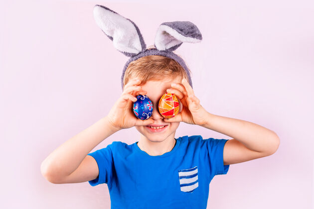 孩子可爱的小男孩头上戴着兔子耳朵 闭上眼睛 粉色背景上有彩色的鸡蛋魅力节日童年