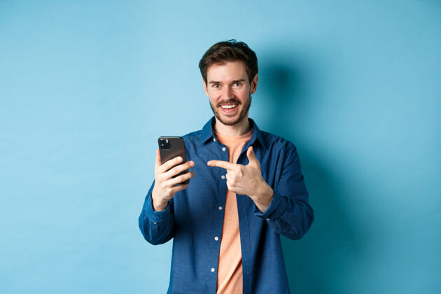 网络帅哥笑眯眯地指着智能手机 高兴地看着相机 推荐应用程序 站在蓝色的背景上年轻社交自信