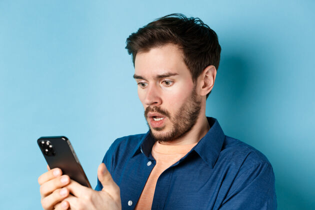 小玩意特写镜头中的男人看起来很震惊 正在智能手机上阅读信息 喘息着 吃惊地盯着屏幕 站在蓝色的背景上时尚胡须科技
