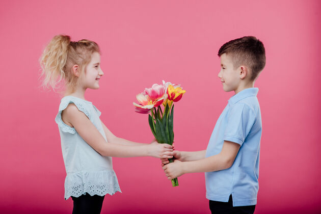 花快乐的小男孩把花伸向一个穿着粉色连衣裙的小女孩 隔离在蓝色的墙上 侧视花束看提议