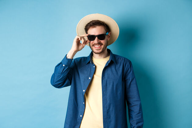 男性快乐微笑的家伙去度暑假 戴着草帽和黑色太阳镜 站在蓝色的背景上成年人眼镜帅哥