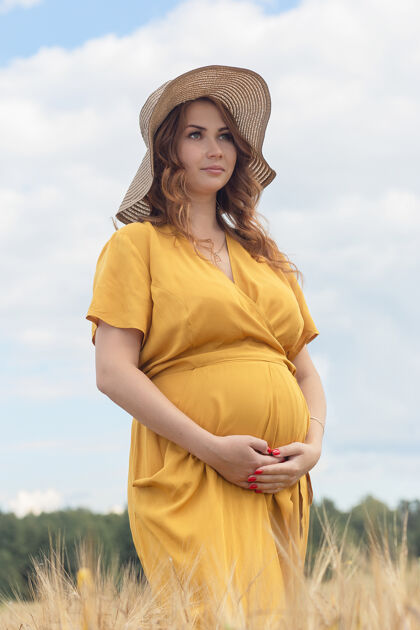 温暖在一个阳光明媚的夏日 一位年轻漂亮的孕妇穿着黄色的裙子 戴着黄色的帽子 走过一片麦田肚子帽子农业