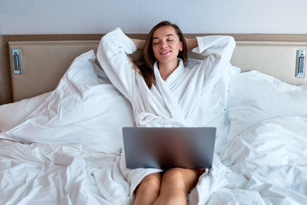 商务女性一位冷静微笑的自由职业女性 双手放在脑后 穿着白色浴袍 在酒店房间的床上电脑旁在线工作轻松的生活方式和满足感女孩博客电脑