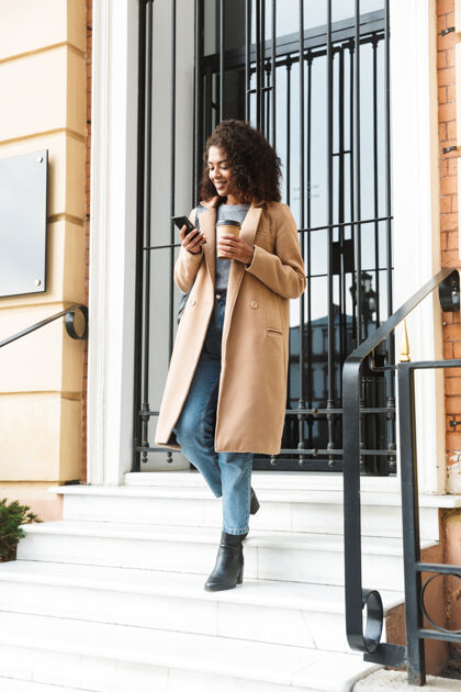 女人快乐的非洲年轻女子穿着外套走在户外 拿着外卖咖啡杯 用手机浏览成人外套