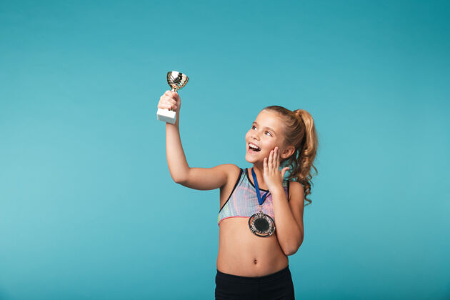 参与快乐的小体育女孩庆祝战胜蓝墙 戴着金牌 展示着奖杯荣誉最佳比赛