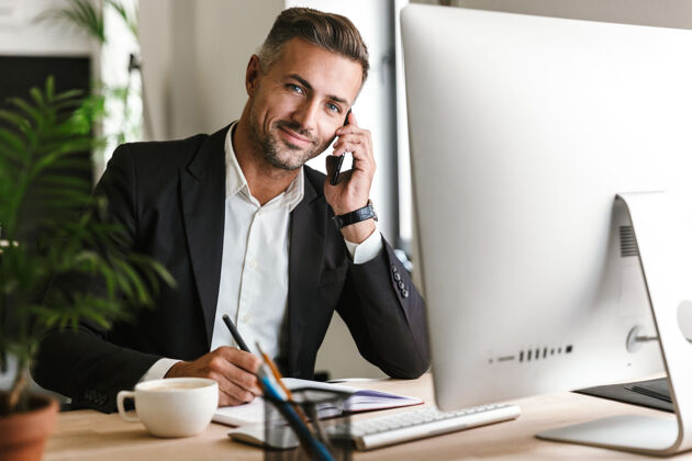 手机30多岁的快乐商人穿着西装在办公室用电脑打电话男士商务型西装