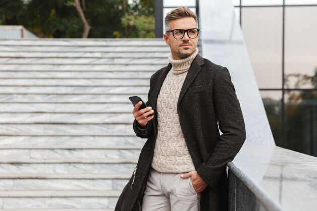 时尚30多岁的成年男子戴着眼镜 穿过城市街道 使用智能手机肖像手机电话