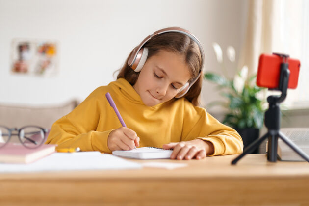 远程教育专心的小女孩在房间里做作业 在家里学习 用带三脚架和耳机的智能手机社会距离家庭教育女学生