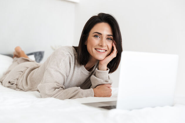 笔记本电脑一张30多岁的快乐女人在明亮的房间里躺在白色亚麻布床上使用笔记本电脑的照片沙发深色头发舒适