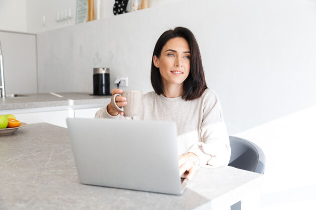 肖像微笑的女人30多岁的工作笔记本电脑 而坐在白色的墙壁在明亮的房间舒适笔记本电脑笔记本电脑