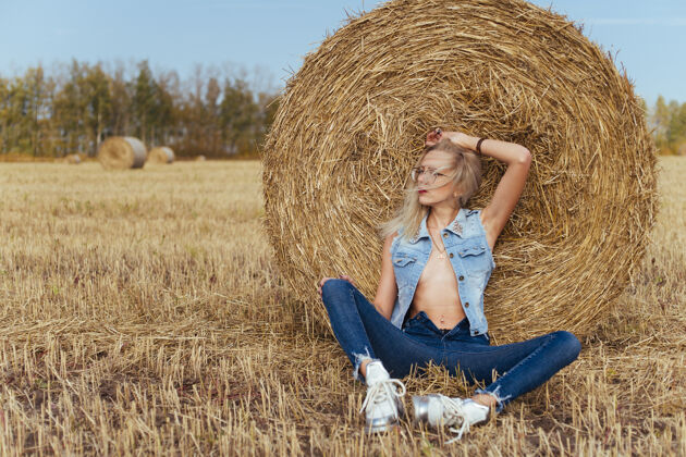 玻璃一位年轻漂亮的女村民穿着牛仔裤在一捆干草旁摆姿势美丽姿势田野