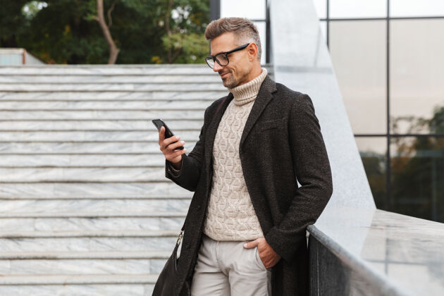 肖像一个30多岁的欧洲男人戴着眼镜 穿过城市街道 用着智能手机户外步行聊天