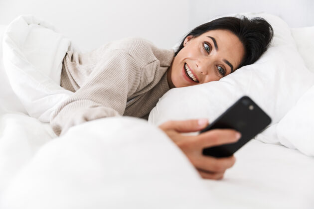 深褐色30多岁的美女在家里穿着白色亚麻布躺在床上用智能手机拍照吸引互动室内