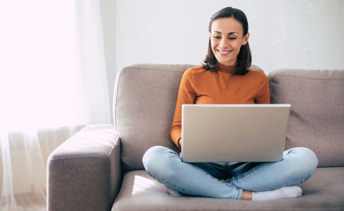 女孩年轻自信的美女坐在家里的沙发上用笔记本电脑工作浏览无线写作