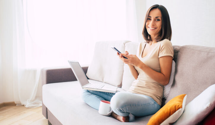 休闲年轻的现代自信微笑的女人在家工作或放松 坐在沙发上用智能手机年轻房间家庭