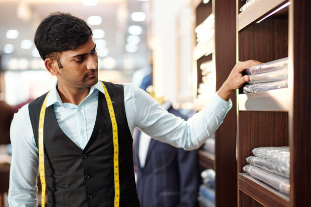服装严肃的印度年轻裁缝看着包装好的成衣 在货架上下单年轻人成人商人