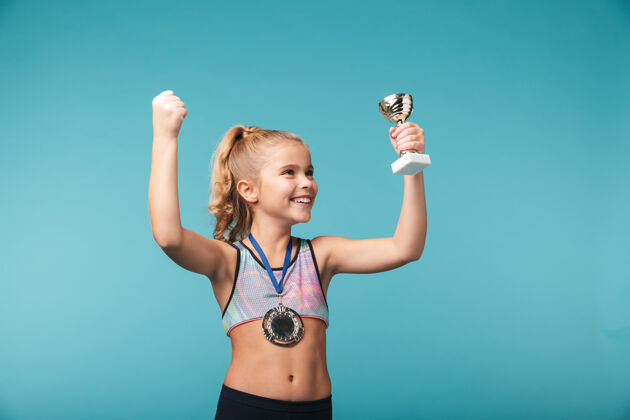 奖杯快乐的小体育女孩庆祝战胜蓝墙 戴着金牌 展示着奖杯成就庆祝比赛