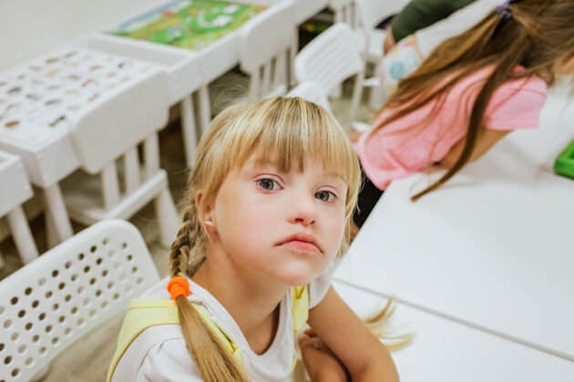 智力患有唐氏综合症的金发女孩的肖像 尾巴和其他孩子坐在白色的书桌上学习医院社交人