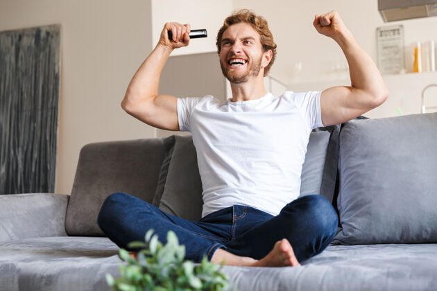 手表照片中 一位帅哥情绪激动地坐在家里的沙发上 拿着遥控器看电视 做出赢家的手势沙发吸引控制