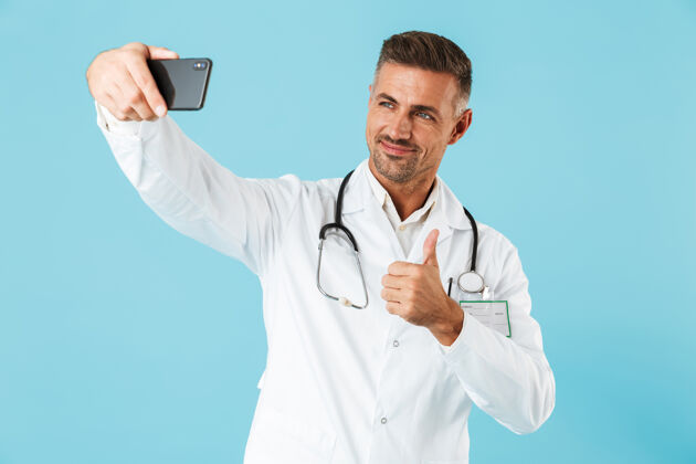 电话身穿白大褂 手持听诊器的年轻医生在智能手机上自拍的照片 孤零零地站在蓝色的墙上中年从业者外套