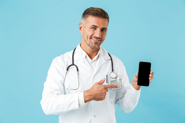 治疗微笑的医生身穿白大褂 手持听诊器手持手机 孤立地站在蓝色的墙上工人听诊器心脏病专家