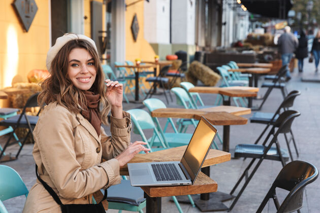 街道穿着外套的漂亮年轻女子坐在咖啡馆外面用笔记本电脑咖啡馆短信秋天
