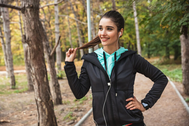 运动员微笑的健身女 戴着耳机听音乐 手持手机站在公园里户外女人听力