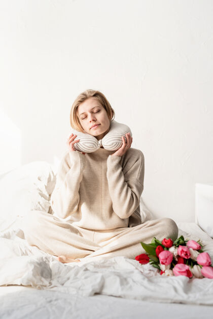 睡眠昏昏欲睡的女人穿着睡衣坐在床上 脖子上套着枕头 床上放着郁金香花微笑节日卧室