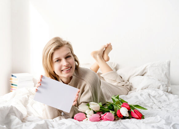 生日快乐的女人穿着睡衣躺在床上 手里拿着郁金香花束和一张设计模型的空白卡片情人节人年轻