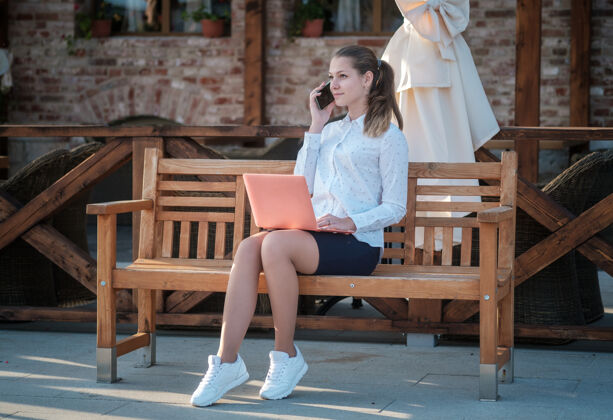 学校一位年轻漂亮的女士坐在长凳上 手里拿着智能手机和笔记本电脑带着笔记本电脑和智能手机的学生在城市街道上学生通信商业